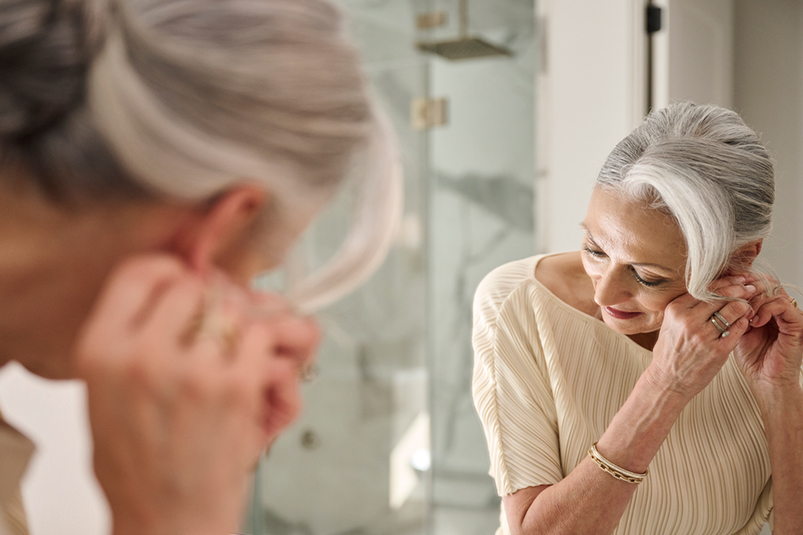 mirror image of a woman in a white dress wearing a bracelet and putting on her earrings